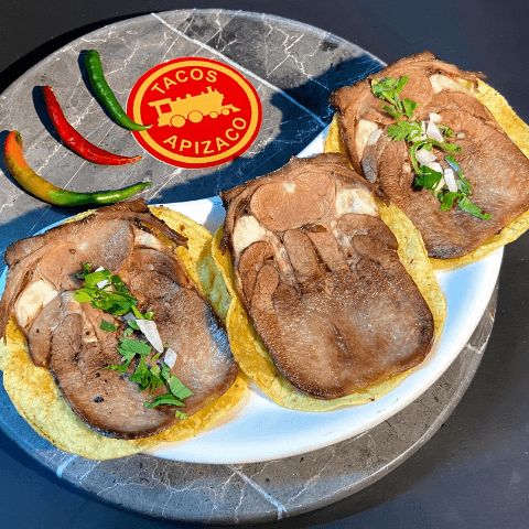 Tres tortillas con rebanadas de lengua de res, cilantro y cebolla, con chiles al lado sobre mármol.